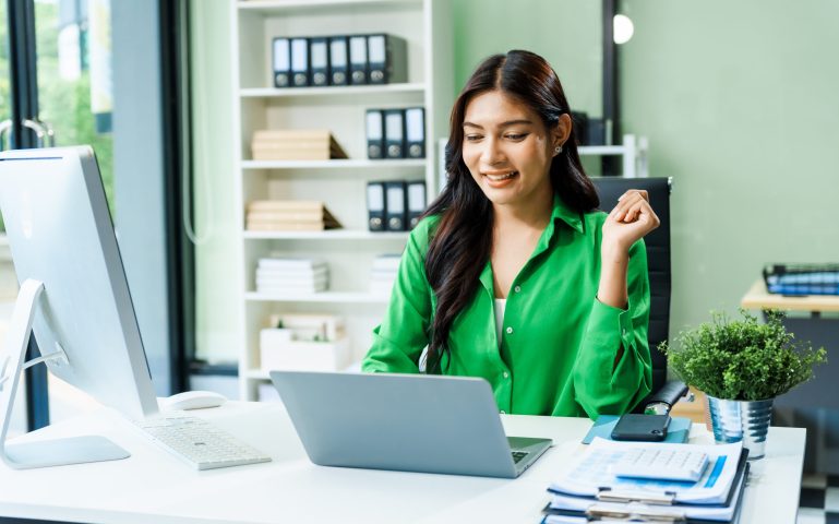 successful and happy young Asian businesswoman in a formal suit works at her desk in a modern glass office. consults on business strategies LGBTQ inclusivity, managing multiple projects efficiently.
